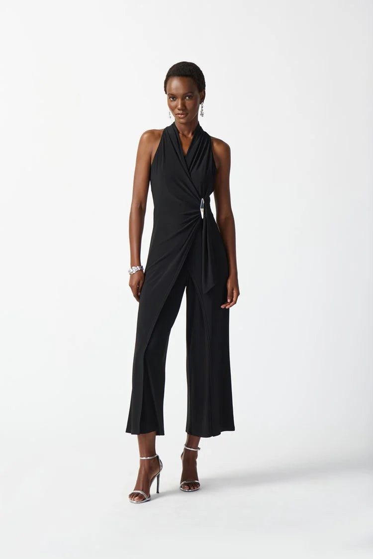 A person with short hair is wearing the Silky Knit Wrap Culotte Jumpsuit 242197 Hero Style by Joseph Ribkoff, a sleeveless, halter-neck jumpsuit that drapes in the front. They have paired it with silver strappy heels and are standing against a white background, smiling and accessorized with a bracelet and earrings.