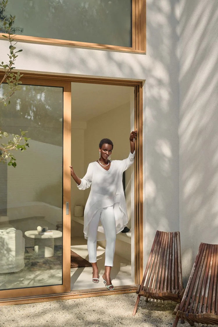 A woman stands against a plain white background wearing the Gauze and Mesh High Low Flare Top 242066 Joseph Ribkoff, a flowy, semi-sheer tunic by Joseph Ribkoff in gauze fabric with three-quarter sleeves that button at the cuffs. She pairs the top with fitted white pants and white open-toe heels, accessorizing with long, dangling earrings.