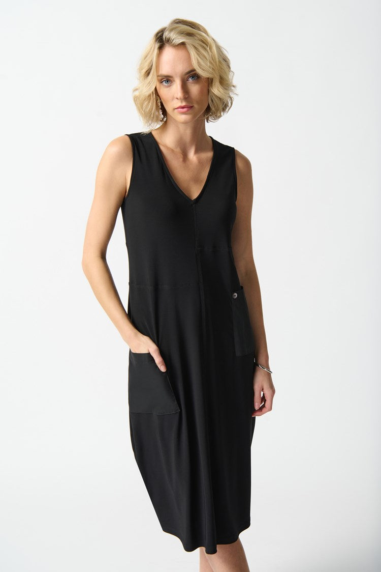 A woman with blonde hair, wearing the Joseph Ribkoff Silky Knit Cocoon Dress with Pockets (style 242161), stands against a plain white background. She has a neutral expression and is posing with one hand in one of the chic, functional front pockets of her V-neckline, sleeveless black dress.