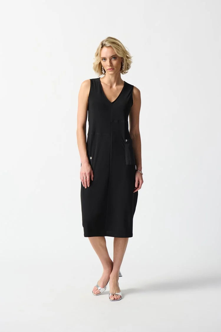 A woman with blonde hair, wearing the Joseph Ribkoff Silky Knit Cocoon Dress with Pockets (style 242161), stands against a plain white background. She has a neutral expression and is posing with one hand in one of the chic, functional front pockets of her V-neckline, sleeveless black dress.