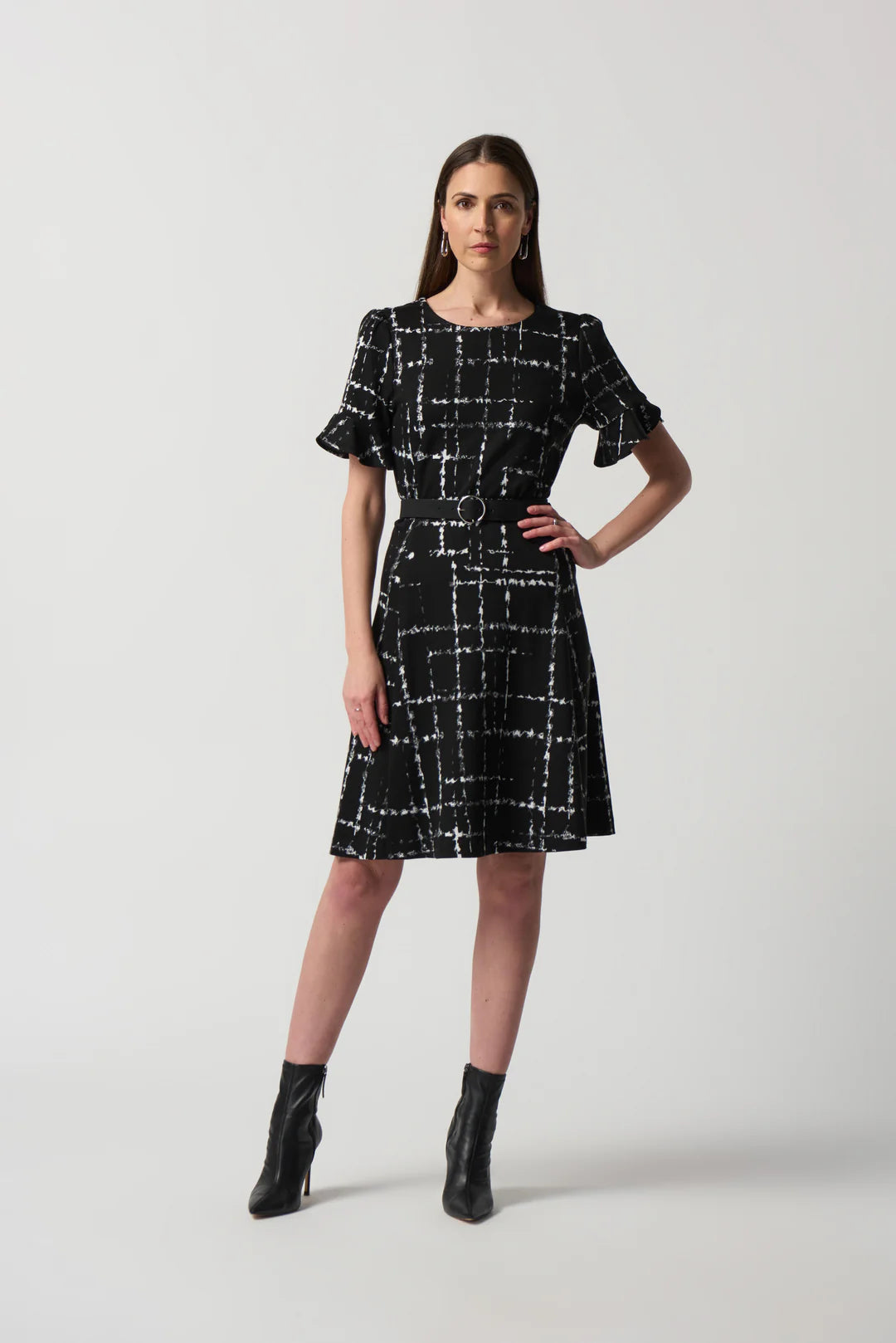 A woman stands confidently against a plain background, wearing the Flutter Sleeve Tweed Dress Style 233004 by Joseph Ribkoff. The dress features a black and white checkered pattern, short ruffled sleeves, and belted detailing around the waist. She pairs it elegantly with black ankle boots.