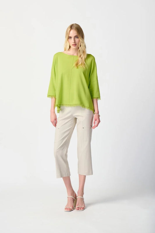 A woman with long blonde hair wears a lime green, loose-fitting top with fringed sleeves and hem, paired with the Joseph Ribkoff Millennium Crop Pull-on Pants (style 241163). She stands against a plain white background, and her white strappy heels complete the outfit.