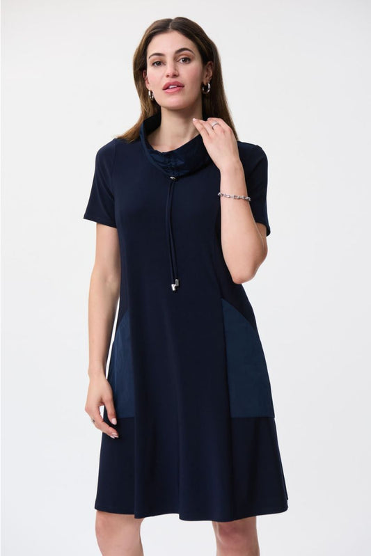 A woman stands against a plain white background, wearing the Drawstring Shirt Dress Style 231141 by Joseph Ribkoff. This navy blue shirt dress is crafted from lightweight fabric and features side pockets and a drawstring collar. She has long, dark hair and is accessorized with subtle jewelry, including a bracelet and earrings. One of her hands rests near her neck.