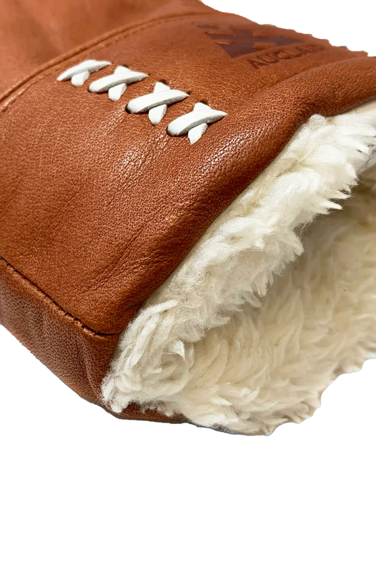 A close-up of the AUCLAIR Anouk Mitts for women showcases the durable brown cow leather with a plush white fur lining at the cuff. White stitching forms an X pattern along the side for added protection. The texture of the leather and the plushness of the fur lining are clearly visible.