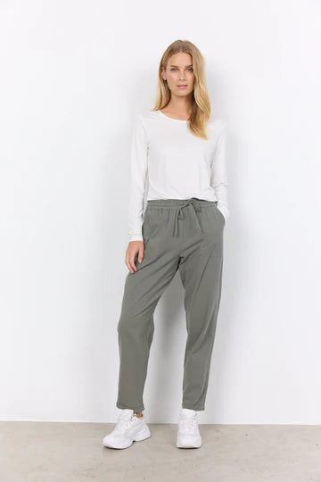 A woman stands against a plain white background wearing a long-sleeved white shirt, SC-CISSIE 10-C pants in Dusty Green by SoyaConcept made of 100% cotton, and white sneakers. She has long, blond hair and her hands are partly in her pockets. The comfortable pants feature a cropped leg length, enhancing the relaxed look.