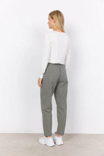 A woman stands against a plain white background wearing a long-sleeved white shirt, SC-CISSIE 10-C pants in Dusty Green by SoyaConcept made of 100% cotton, and white sneakers. She has long, blond hair and her hands are partly in her pockets. The comfortable pants feature a cropped leg length, enhancing the relaxed look.