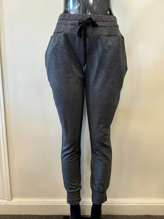 The Cosy Up Tie Jogger by FOIL is showcased on a black mannequin. These gray joggers come with an elastic waistband and a black drawstring for a customizable fit, along with convenient side pockets. The fabric looks soft and textured, making it ideal for any occasion. The mannequin is positioned against a cream-colored wall.