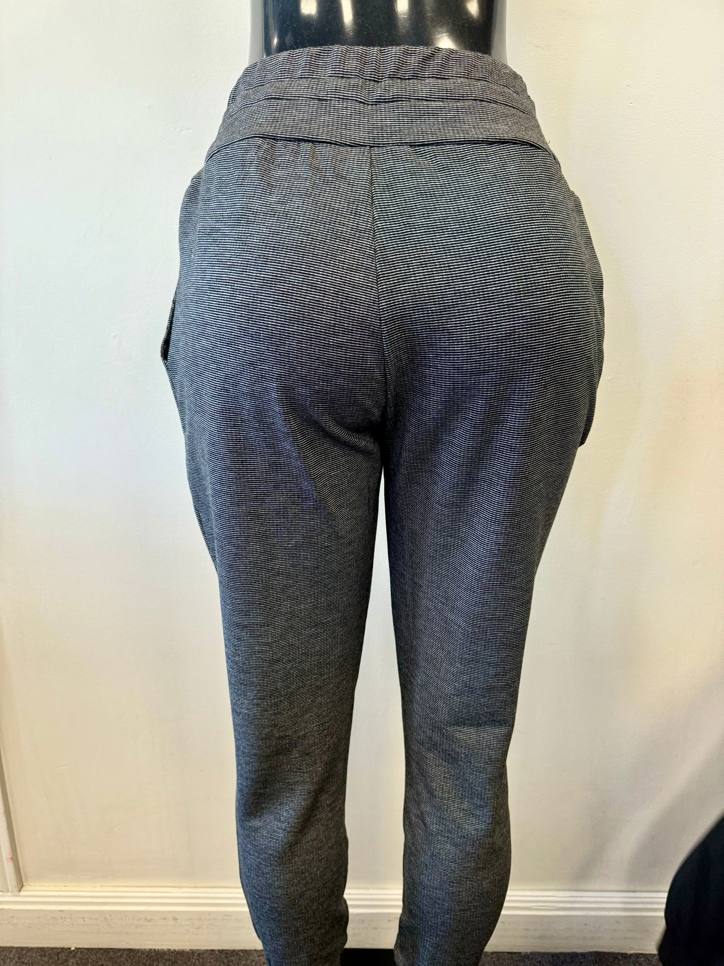 A mannequin is dressed in FOIL's Cosy Up Tie Jogger, featuring light grey joggers with a black drawstring at the waist for a customizable fit. These versatile pants are made of knit fabric, have pockets, and feature cuffed ankles. The background is a plain off-white wall.