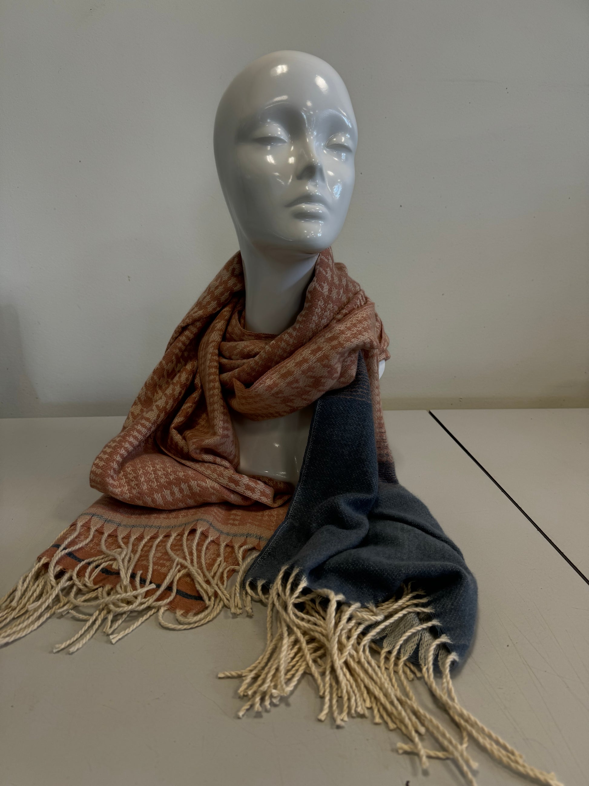 A white display mannequin with a smooth head is adorned with the luxurious Majestic Cashmere Scarf by Catherine Lillywhite. The scarf is wrapped around the neck, featuring an elegant color combination that transitions from a light red checkered pattern on one end to a solid dark grayish-blue color with fringed edges on the other.