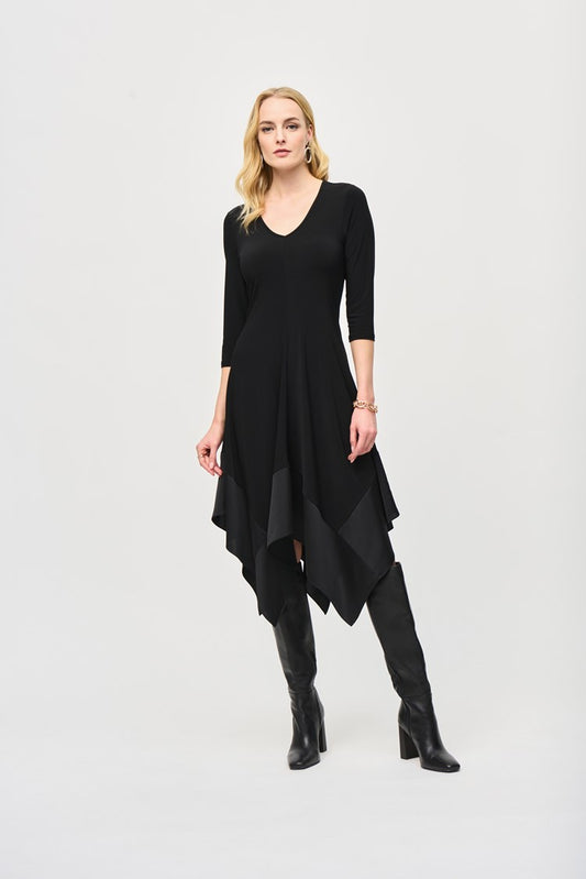 A woman stands confidently against a plain background wearing the Joseph Ribkoff Silky Knit Handkerchief Dress (243092), featuring an asymmetrical hemline, a V-shaped neckline, and three-quarter sleeves. She has long, blonde hair and is wearing black knee-high boots. Her hands rest by her sides, and she has a composed expression.