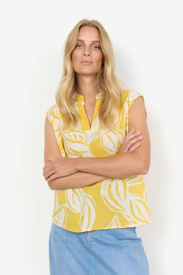 A woman with long blonde hair stands with arms crossed, wearing the SC-ELVINE 1 TOP YELLOW by SoyaConcept—a sleeveless, feminine top made of organic cotton featuring a white leaf pattern—and a light blue denim skirt. She faces the camera against a white background.