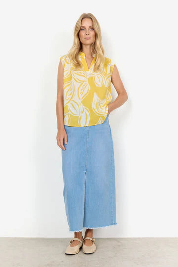 A woman with long blonde hair stands with arms crossed, wearing the SC-ELVINE 1 TOP YELLOW by SoyaConcept—a sleeveless, feminine top made of organic cotton featuring a white leaf pattern—and a light blue denim skirt. She faces the camera against a white background.