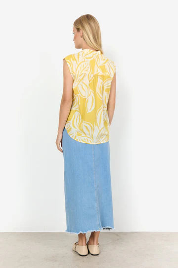 A woman with long blonde hair stands with arms crossed, wearing the SC-ELVINE 1 TOP YELLOW by SoyaConcept—a sleeveless, feminine top made of organic cotton featuring a white leaf pattern—and a light blue denim skirt. She faces the camera against a white background.