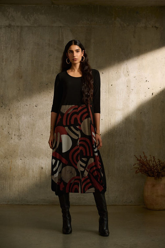 A person with long dark hair stands against a concrete wall, bathed in a streak of sunlight. They are wearing the Silky Knit Geometric Print Cocoon Dress 243234 by Joseph Ribkoff, featuring red and beige circular designs. Black boots complete the outfit. A vase with dried flowers is on the floor beside them.