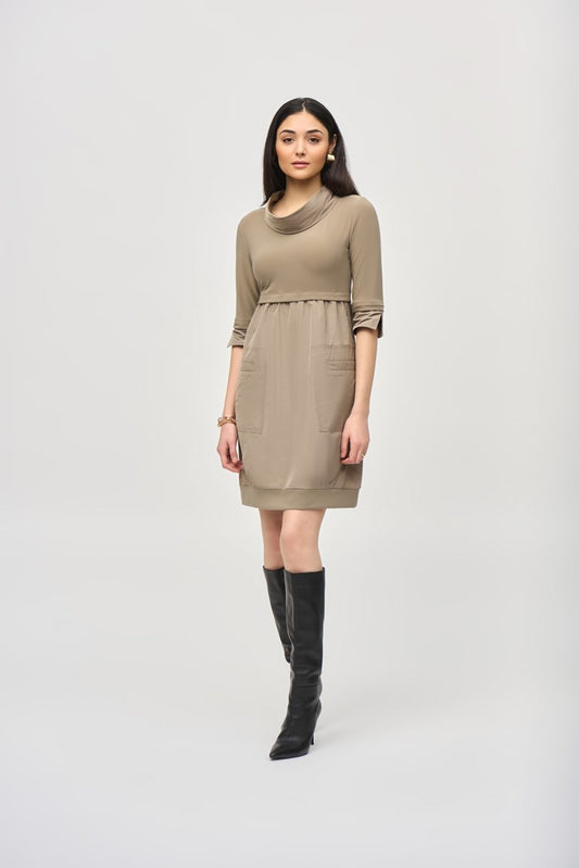 A woman with long dark hair stands against a plain white background, wearing the Cocoon Dress - Seasonal Colors 173444F24 by Joseph Ribkoff. The taupe, long-sleeve, knee-length dress features a cowl neckline and convenient pockets. She pairs it with black knee-high boots and has a neutral expression on her face.