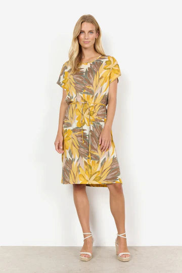 A woman stands against a plain white background wearing the SC-FELICITY AOP 457 DRESS YELLOW by SoyaConcept, a short-sleeve, knee-length dress featuring a floral print with yellow, brown, and green leaf patterns. She has long blonde hair and is wearing white high-heeled sandals. Her hands are at her sides as she looks slightly to the left.