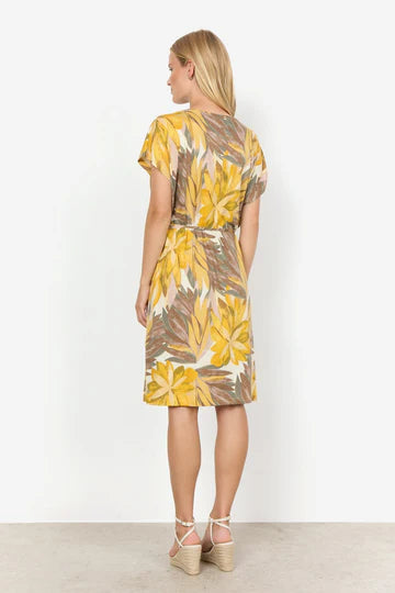 A woman stands against a plain white background wearing the SC-FELICITY AOP 457 DRESS YELLOW by SoyaConcept, a short-sleeve, knee-length dress featuring a floral print with yellow, brown, and green leaf patterns. She has long blonde hair and is wearing white high-heeled sandals. Her hands are at her sides as she looks slightly to the left.