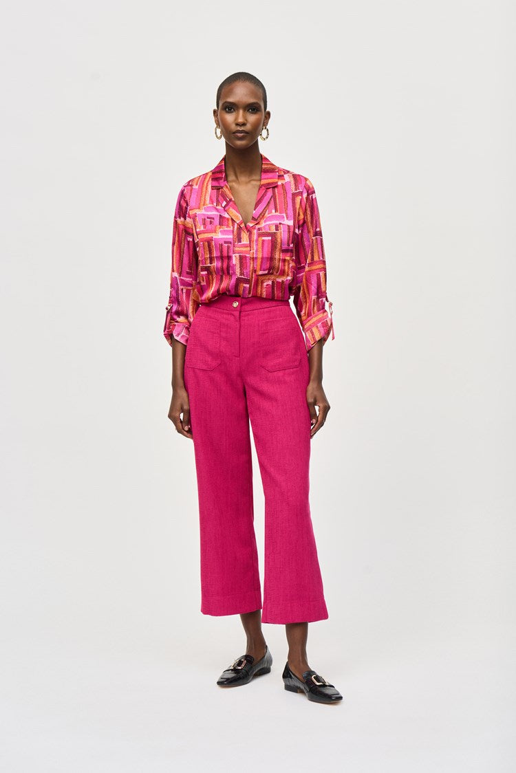 A person is shown wearing Joseph Ribkoff's Corduroy Culotte Pant 243295 in a vibrant pink jacquard pattern, featuring visible front pockets and a button closure. The top appears to be a colorful blouse with shades of pink, orange, and white. One hand is casually placed into a pocket.