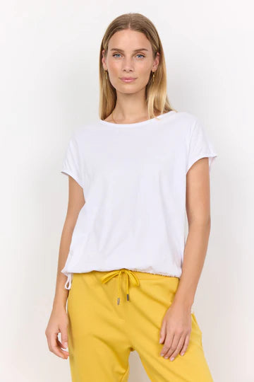 A person with long, blonde hair is wearing a white SC-DERBY 33 T-SHIRT from SoyaConcept, featuring a round neckline. They have paired it with yellow drawstring pants and are standing against a plain, light-colored background. Their expression is neutral as they look directly at the camera.