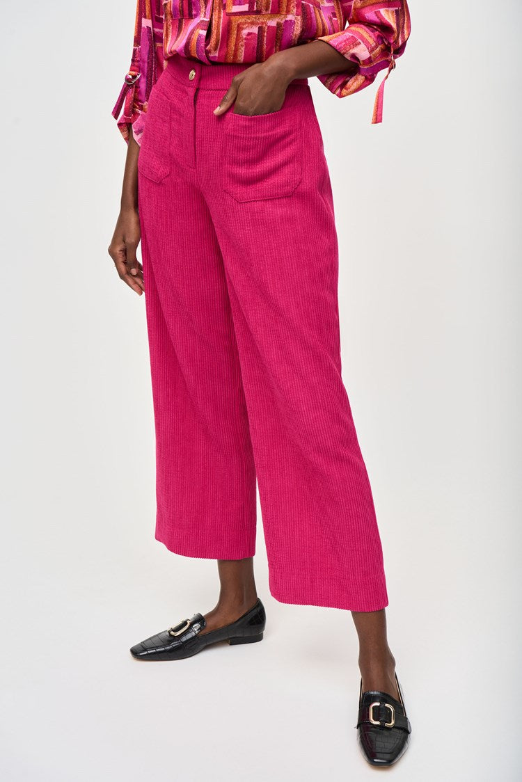 A person is shown wearing Joseph Ribkoff's Corduroy Culotte Pant 243295 in a vibrant pink jacquard pattern, featuring visible front pockets and a button closure. The top appears to be a colorful blouse with shades of pink, orange, and white. One hand is casually placed into a pocket.