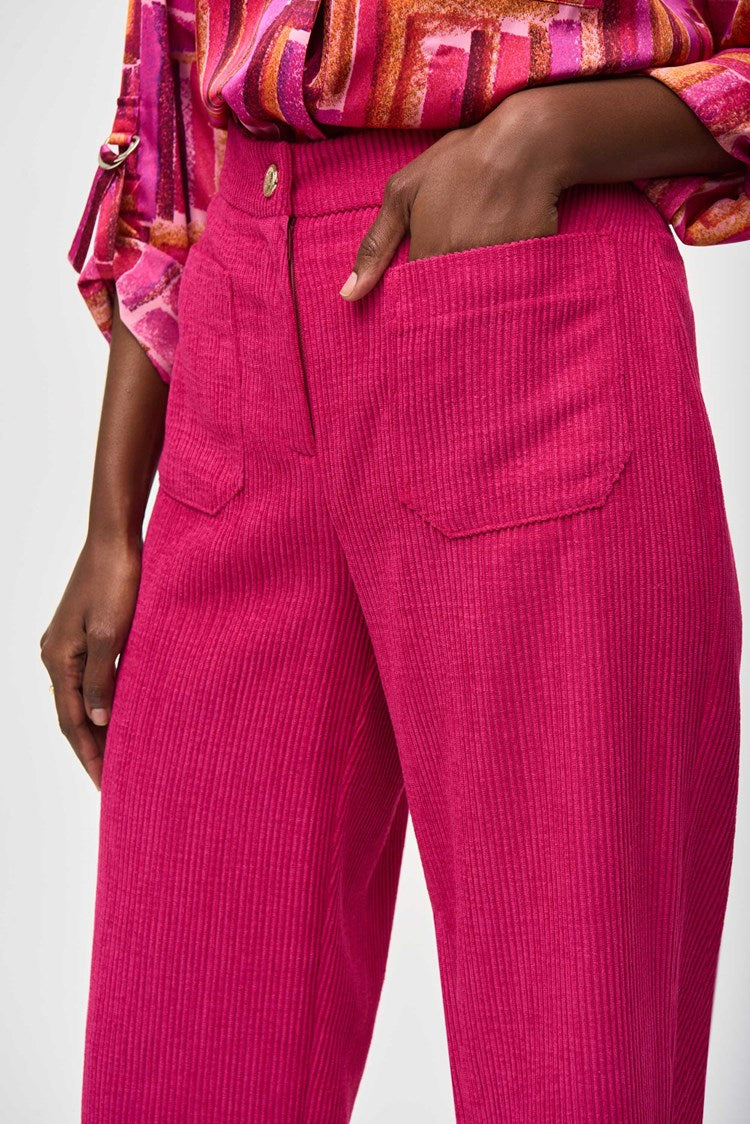 A person is shown wearing Joseph Ribkoff's Corduroy Culotte Pant 243295 in a vibrant pink jacquard pattern, featuring visible front pockets and a button closure. The top appears to be a colorful blouse with shades of pink, orange, and white. One hand is casually placed into a pocket.