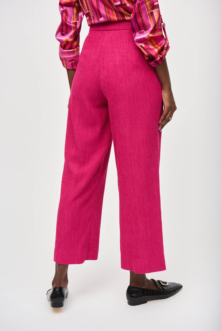 A person is shown wearing Joseph Ribkoff's Corduroy Culotte Pant 243295 in a vibrant pink jacquard pattern, featuring visible front pockets and a button closure. The top appears to be a colorful blouse with shades of pink, orange, and white. One hand is casually placed into a pocket.