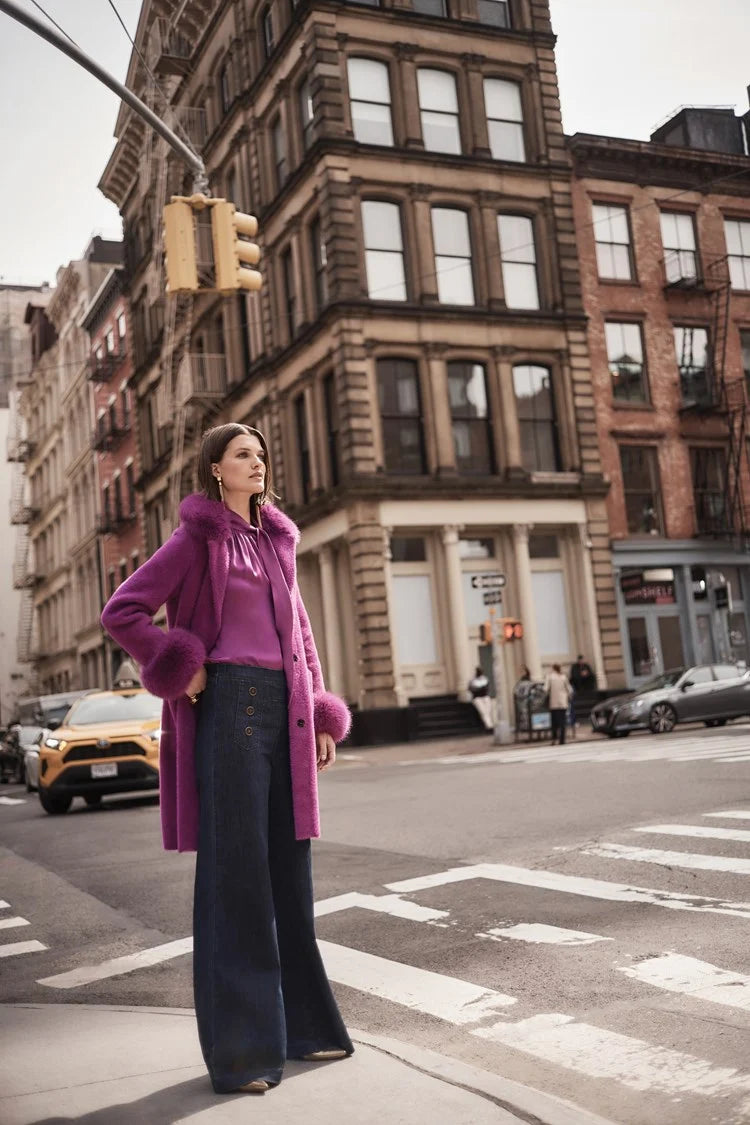 A woman stands against a plain background wearing a green long-sleeve blouse with a V-neckline and dark blue High-Rise Wide Leg Denim Pants 243900 by Joseph Ribkoff adorned with gold buttons. She completes her retro look with white pointed-toe heels and large, round earrings.