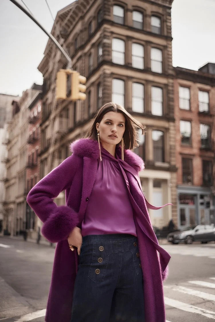 A woman stands against a plain background wearing a green long-sleeve blouse with a V-neckline and dark blue High-Rise Wide Leg Denim Pants 243900 by Joseph Ribkoff adorned with gold buttons. She completes her retro look with white pointed-toe heels and large, round earrings.