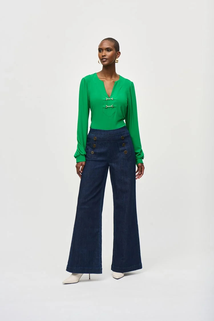 A woman stands against a plain background wearing a green long-sleeve blouse with a V-neckline and dark blue High-Rise Wide Leg Denim Pants 243900 by Joseph Ribkoff adorned with gold buttons. She completes her retro look with white pointed-toe heels and large, round earrings.