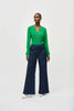 A woman stands against a plain background wearing a green long-sleeve blouse with a V-neckline and dark blue High-Rise Wide Leg Denim Pants 243900 by Joseph Ribkoff adorned with gold buttons. She completes her retro look with white pointed-toe heels and large, round earrings.