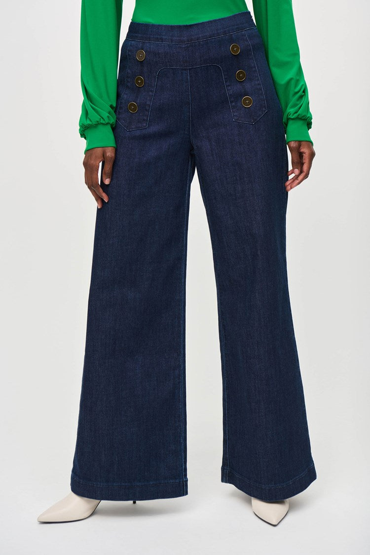 A woman stands against a plain background wearing a green long-sleeve blouse with a V-neckline and dark blue High-Rise Wide Leg Denim Pants 243900 by Joseph Ribkoff adorned with gold buttons. She completes her retro look with white pointed-toe heels and large, round earrings.