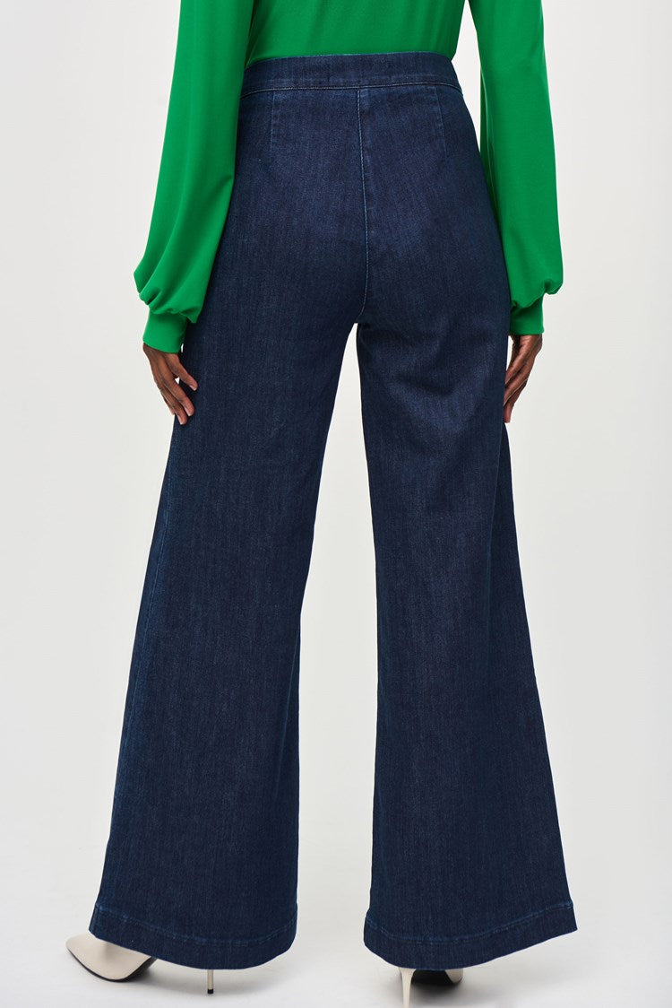 A woman stands against a plain background wearing a green long-sleeve blouse with a V-neckline and dark blue High-Rise Wide Leg Denim Pants 243900 by Joseph Ribkoff adorned with gold buttons. She completes her retro look with white pointed-toe heels and large, round earrings.