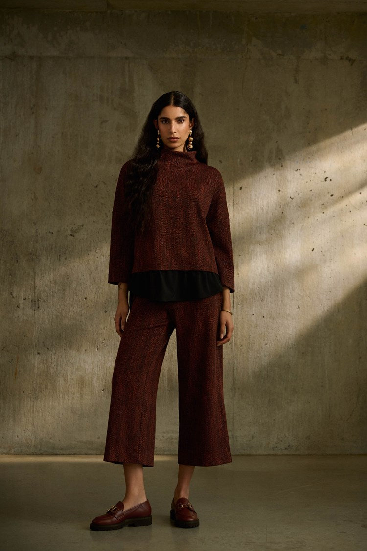 A person with long dark hair stands in front of a textured concrete wall. They are wearing an oversized maroon top and matching Joseph Ribkoff Jacquard Knit Pull-On Culotte Pants 243302 with a black shirt underneath. The outfit is complemented by maroon loafers and gold earrings.