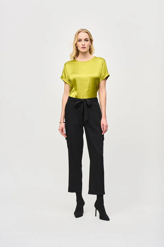 A woman stands against a plain white background, wearing a short-sleeve, lime green blouse paired with the Stretch Woven Pull-On Cargo Pants 243077 by Joseph Ribkoff. The high-waisted, black cropped pants feature a tied belt at the waist. She completes her look with black ankle boots. Her blonde hair is styled in loose waves and she has neutral makeup.
