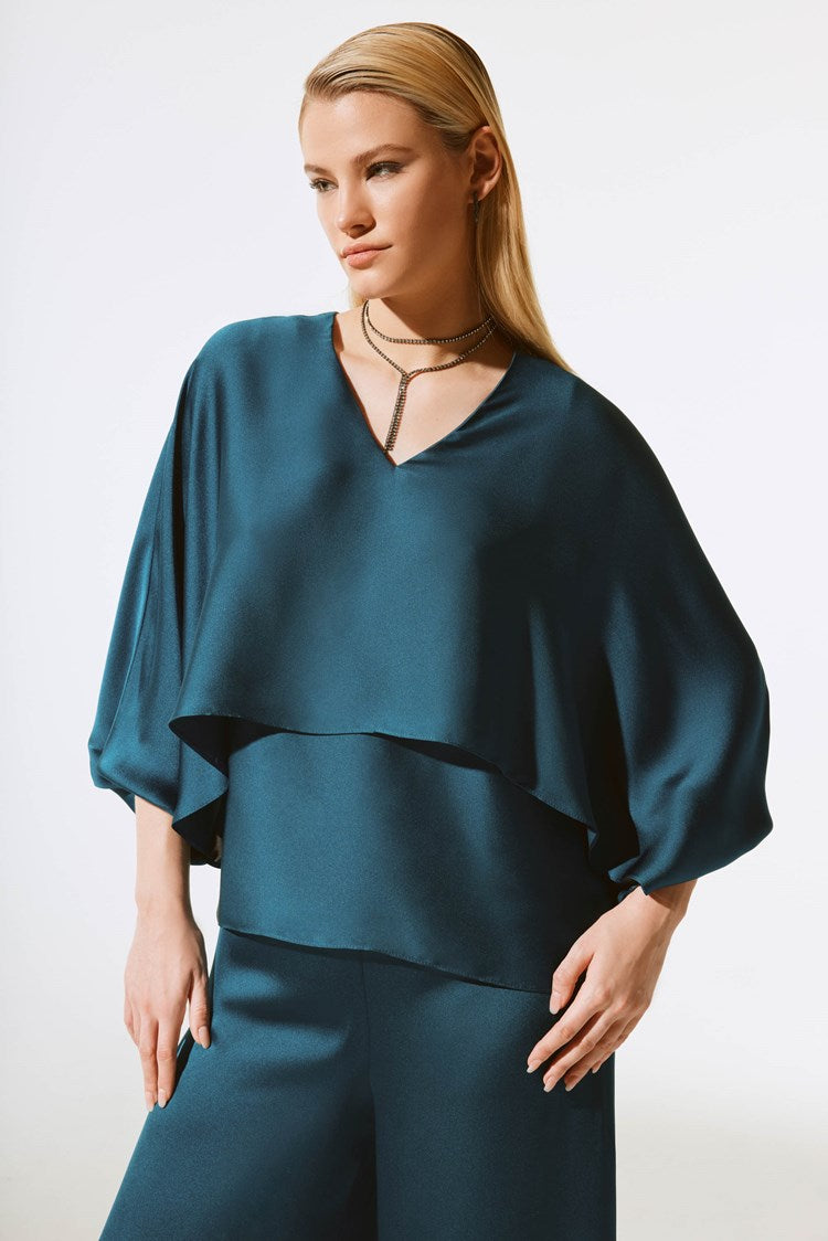 A person with long, straight blonde hair is wearing a teal, loose-fitting Satin V-Neck Layered Top 243790 by Joseph Ribkoff paired with matching pants. They accessorize with a multi-strand necklace featuring a pendant. The background is a plain white studio setting.