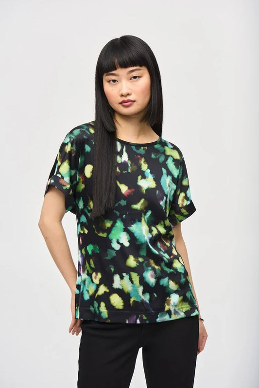 A woman with long, straight black hair and blunt bangs is standing against a plain background. She is wearing the Satin Front Abstract Print Short Sleeve Top (item 243938) by Joseph Ribkoff, which features a knit fabric with a black base adorned with vibrant abstract patterns in green, yellow, and blue hues. She has a neutral expression.