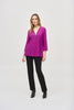 A woman with long hair is standing against a plain background, wearing the Silky Knit Fit and Flare Tunic 243314 by Joseph Ribkoff. The tunic features a long-sleeved, purple V-neck design made of silky knit fabric and adorned with a Rhinestone zipper neckline. She pairs it with black pants and black boots, showcasing a relaxed and confident posture.
