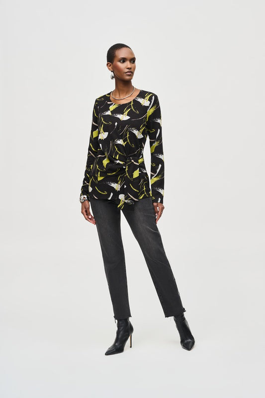 A person stands confidently against a plain white background, wearing a Joseph Ribkoff Sweater Knit Abstract Print Top 243197 featuring a black long-sleeve design with yellow and white abstract patterns and front draping. They pair it with black pants and black heeled boots. Their short hair is styled to highlight the top's scoop neckline, accessorized with earrings, striking a poised pose.