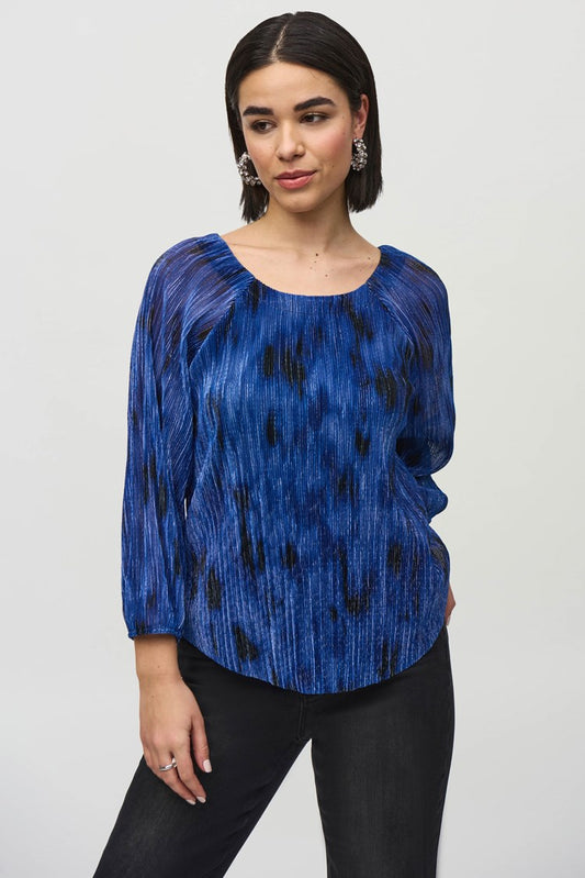 A person with shoulder-length black hair is standing against a plain background, wearing the Joseph Ribkoff Pleated Knit Abstract Print Flared Top 244105, which is blue with long sleeves and features a textured, pleated design. They are also wearing dark pants and are looking slightly to the side with a calm expression.