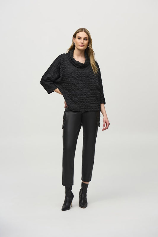 A woman stands against a plain white background, wearing a black Woven Jacquard Boxy Top 244069 by Joseph Ribkoff with three-quarter sleeves, black leather pants with side pockets, and black heeled ankle boots. Her left hand is on her hip and her right arm hangs by her side. She has long, wavy hair.