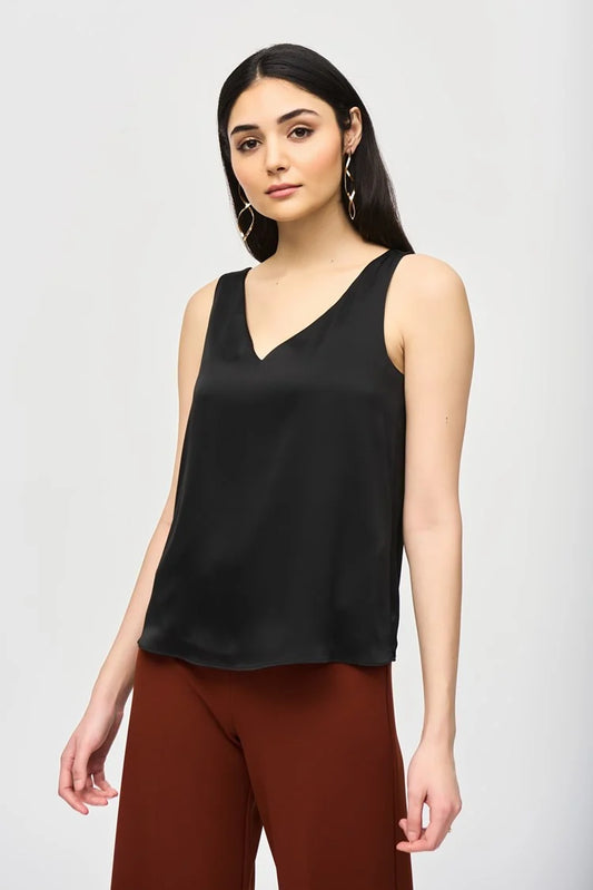 A woman with long, dark hair is wearing a Joseph Ribkoff Satin Straight Sleeveless Top 243110, which features a V-shaped neckline, paired with brown pants. She stands against a plain white background and has a neutral expression. She is also adorned with long, dangling earrings.