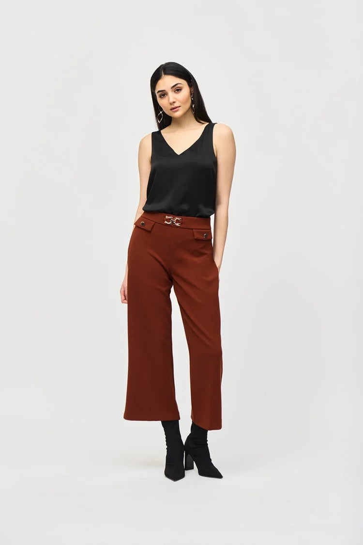 A woman with long, dark hair is wearing a Joseph Ribkoff Satin Straight Sleeveless Top 243110, which features a V-shaped neckline, paired with brown pants. She stands against a plain white background and has a neutral expression. She is also adorned with long, dangling earrings.