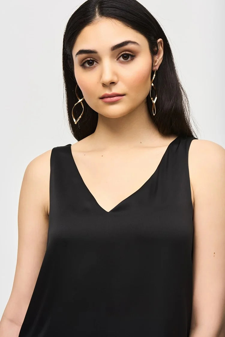 A woman with long, dark hair is wearing a Joseph Ribkoff Satin Straight Sleeveless Top 243110, which features a V-shaped neckline, paired with brown pants. She stands against a plain white background and has a neutral expression. She is also adorned with long, dangling earrings.