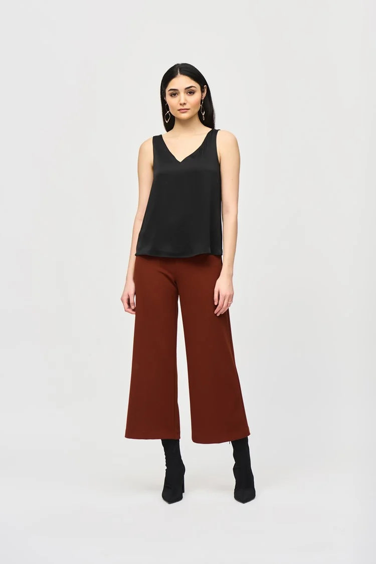 A woman with long, dark hair is wearing a Joseph Ribkoff Satin Straight Sleeveless Top 243110, which features a V-shaped neckline, paired with brown pants. She stands against a plain white background and has a neutral expression. She is also adorned with long, dangling earrings.