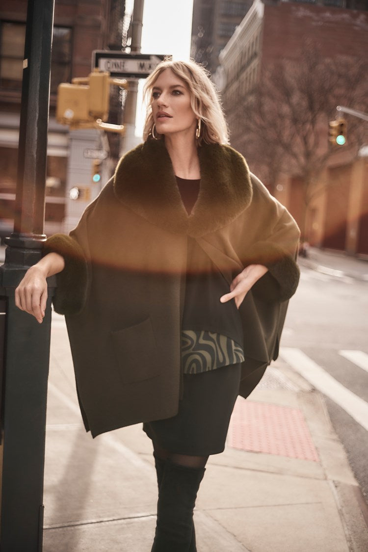 A woman with long dark hair stands against a plain light gray background, wearing the Silky Knit Tunic With Abstract Print Layer from Joseph Ribkoff. She pairs the abstract print black long-sleeve top that features pocket accents with black leather pants and black ankle boots. She looks at the camera with a neutral expression.