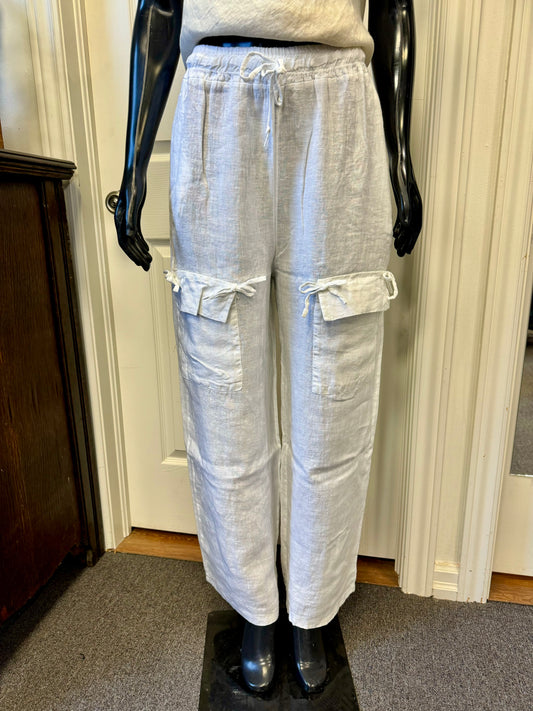 A pair of stylish white ETERNELLE linen pants with pockets, designed by the renowned brand ETERNELLE and named "LINEN PANT WITH POCKET WHITE-ETERNELLEE," is displayed on a black mannequin in a room with light-colored walls and carpet. Made in Italy, these cargo pants feature multiple pockets with drawstring ties and a relaxed fit.