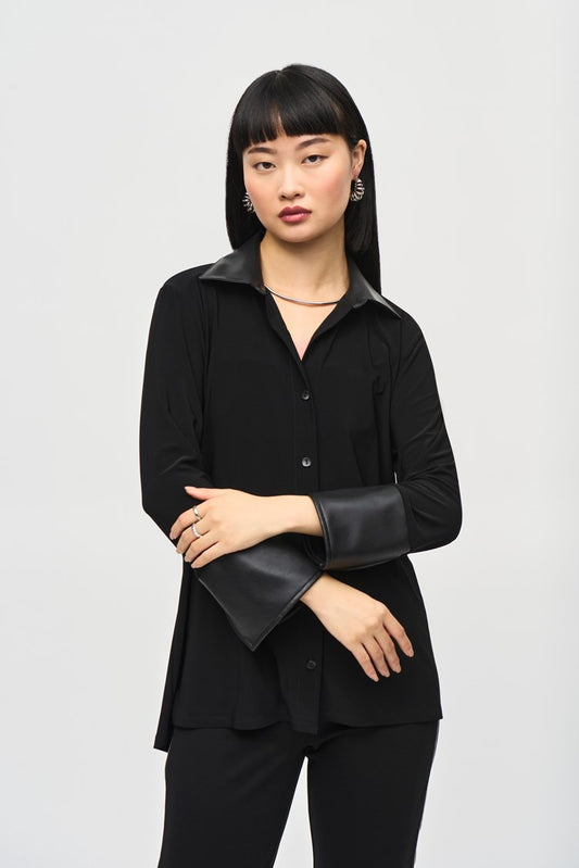 A person with long black hair and bangs poses against a plain background, wearing the Silky Knit Fit and Flare Blouse 243204 by Joseph Ribkoff. The blouse features a leatherette faux collar and leather cuffs, paired elegantly with black pants. Their expression is neutral, and one hand rests gently on their opposite arm, just above the elbow.