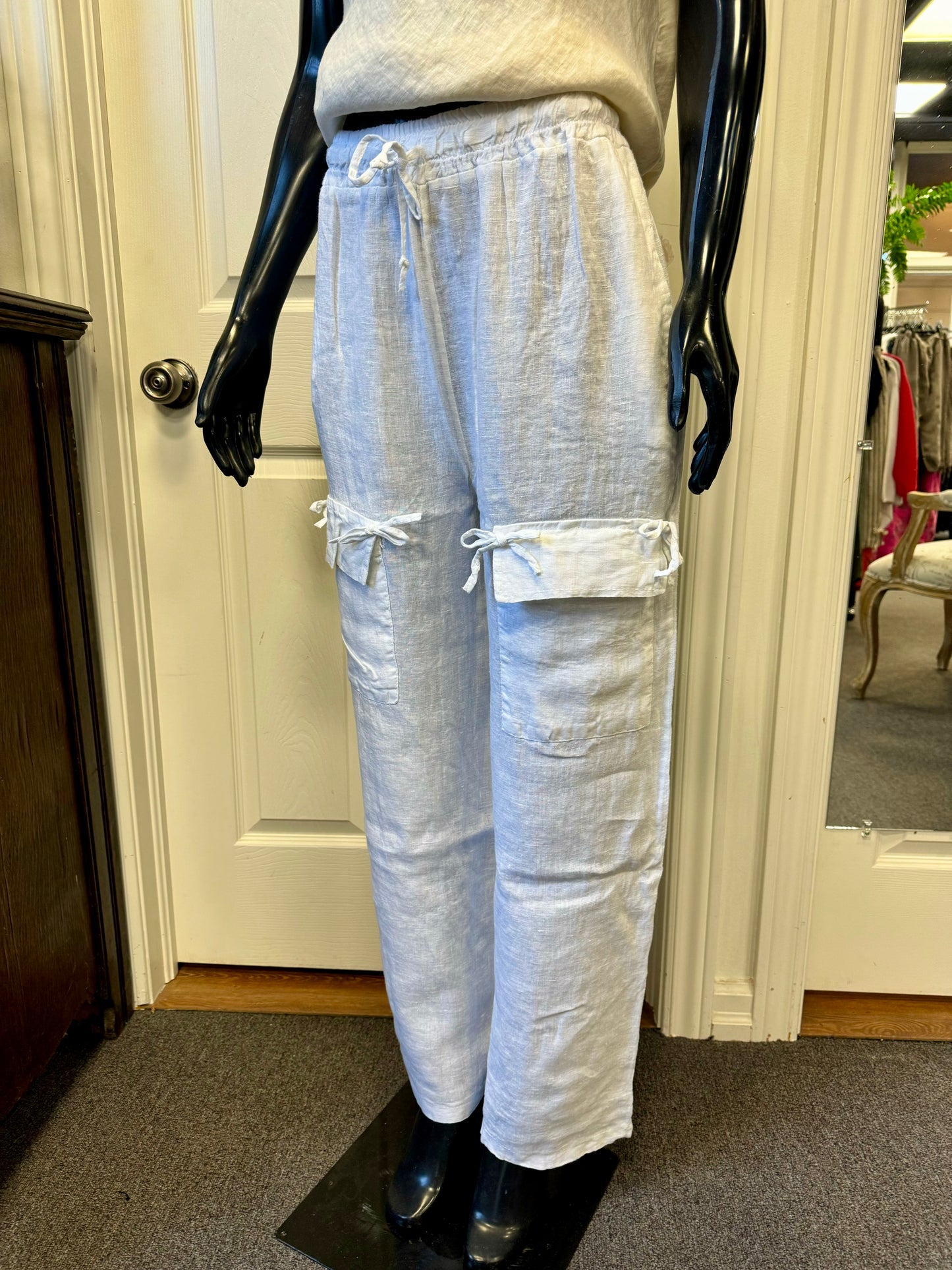 A pair of stylish white ETERNELLE linen pants with pockets, designed by the renowned brand ETERNELLE and named "LINEN PANT WITH POCKET WHITE-ETERNELLEE," is displayed on a black mannequin in a room with light-colored walls and carpet. Made in Italy, these cargo pants feature multiple pockets with drawstring ties and a relaxed fit.