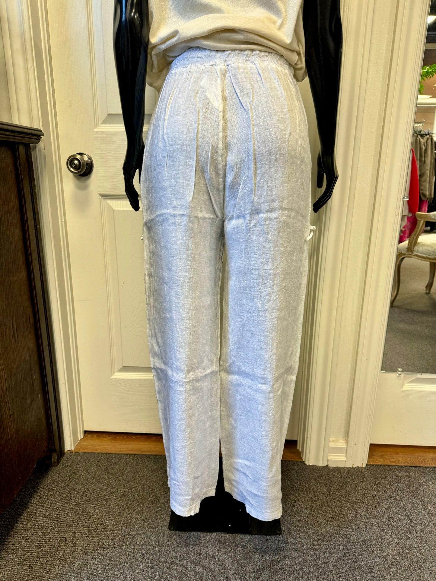 A pair of stylish white ETERNELLE linen pants with pockets, designed by the renowned brand ETERNELLE and named "LINEN PANT WITH POCKET WHITE-ETERNELLEE," is displayed on a black mannequin in a room with light-colored walls and carpet. Made in Italy, these cargo pants feature multiple pockets with drawstring ties and a relaxed fit.