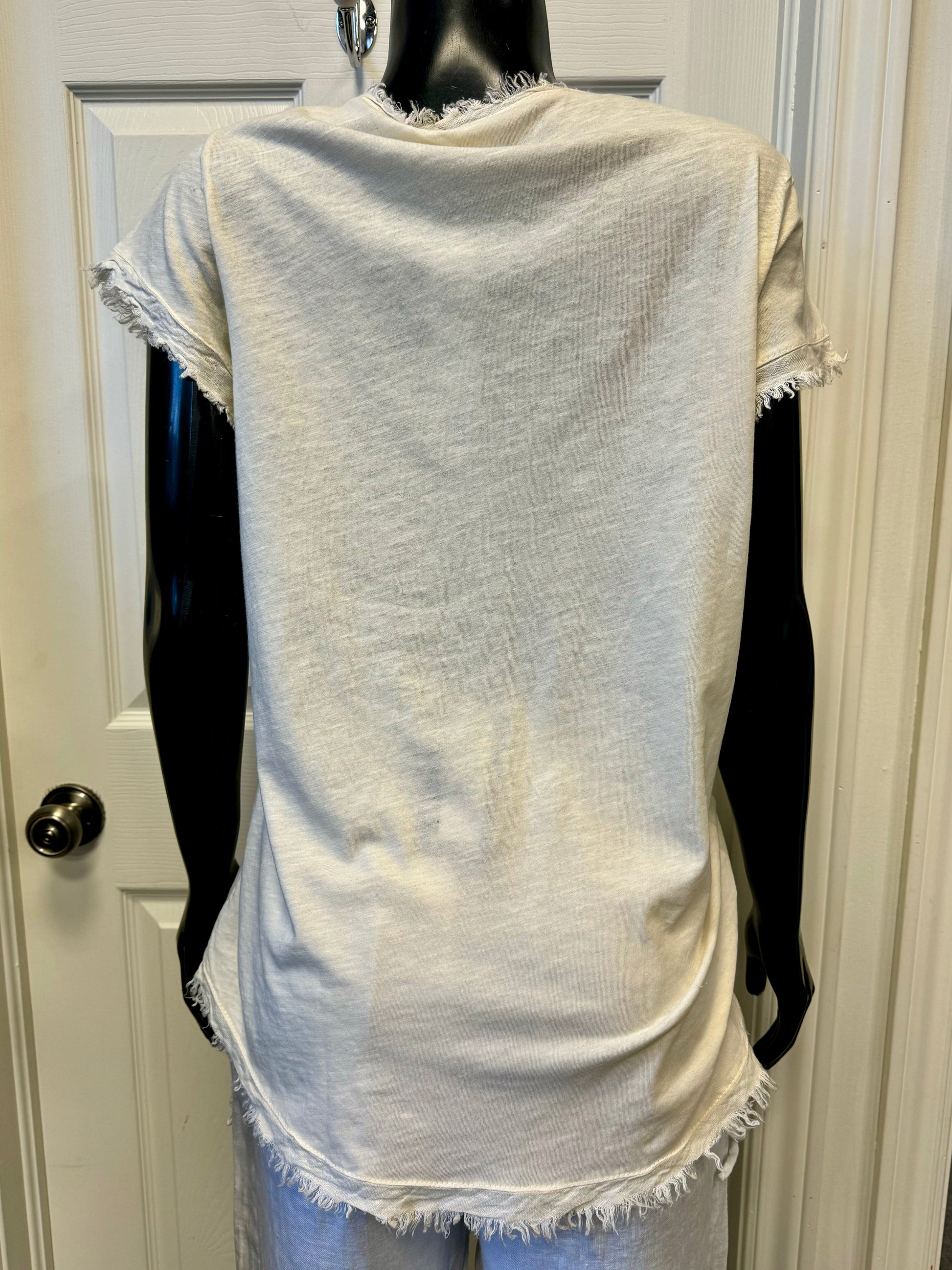 A mannequin showcases the LINEN TOP VANILLA- ETERNELLE by ETERNELLE, featuring a light beige, short-sleeved, V-neck design with frayed edges. The 100% linen fabric has a textured, woven appearance. The vanilla-hued top is paired with matching pants and displayed against a white-paneled door background.